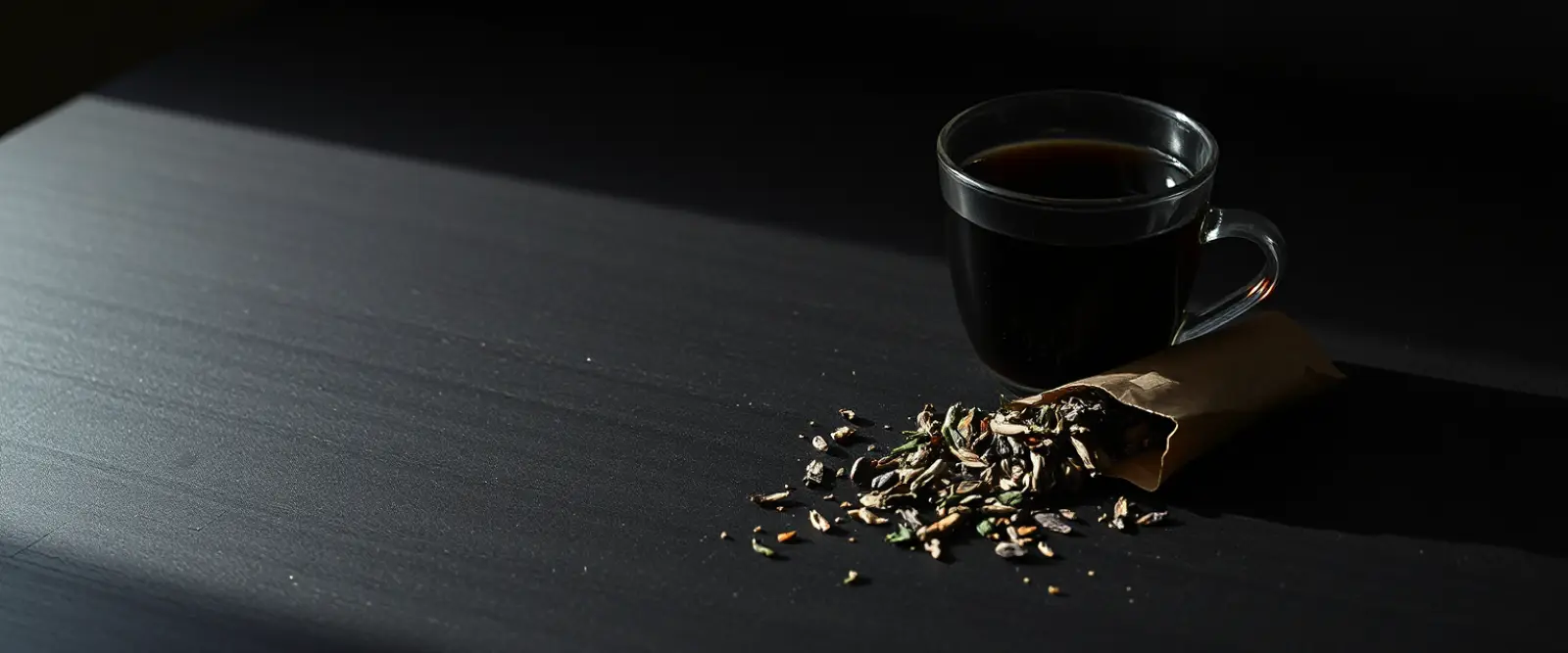 An open paper packet of loose-leaf tea spilling onto a dark table, a glass of brewed tea behind it.