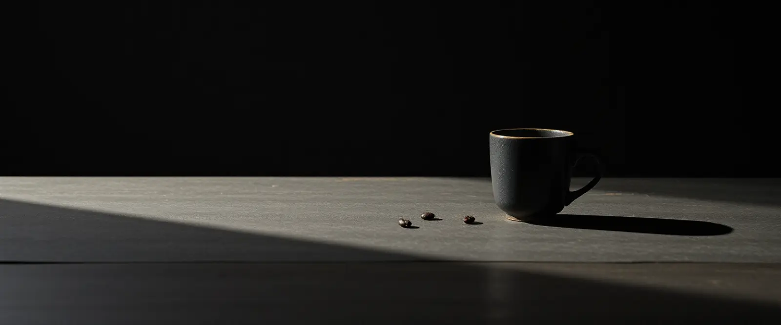 A single dark ceramic cup of black coffee on a wide table in a dim room.