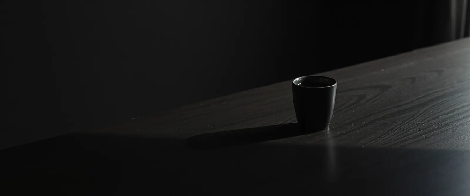 A single dark ceramic cup of black coffee on a wide table in a dim room.