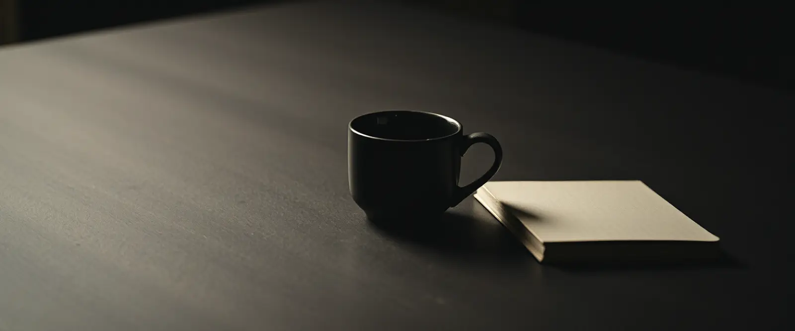 An untouched cup of coffee at the edge of a wide empty table beside a closed notebook.
