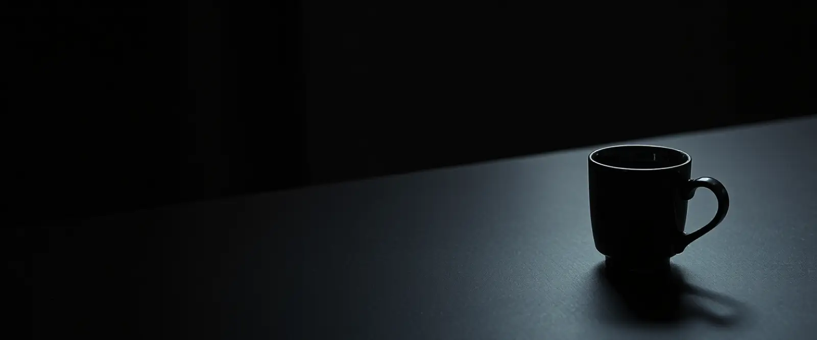 A single dark ceramic cup of black coffee on a wide table in a dim room.