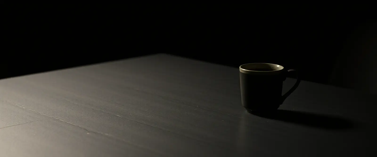 A single dark ceramic cup of black coffee on a wide table in a dim room.