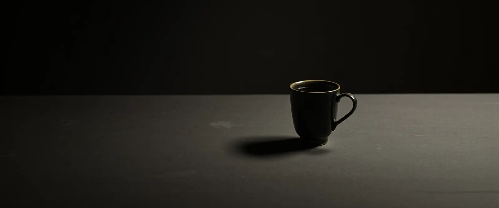 A single dark ceramic cup of black coffee on a wide table in a dim room.