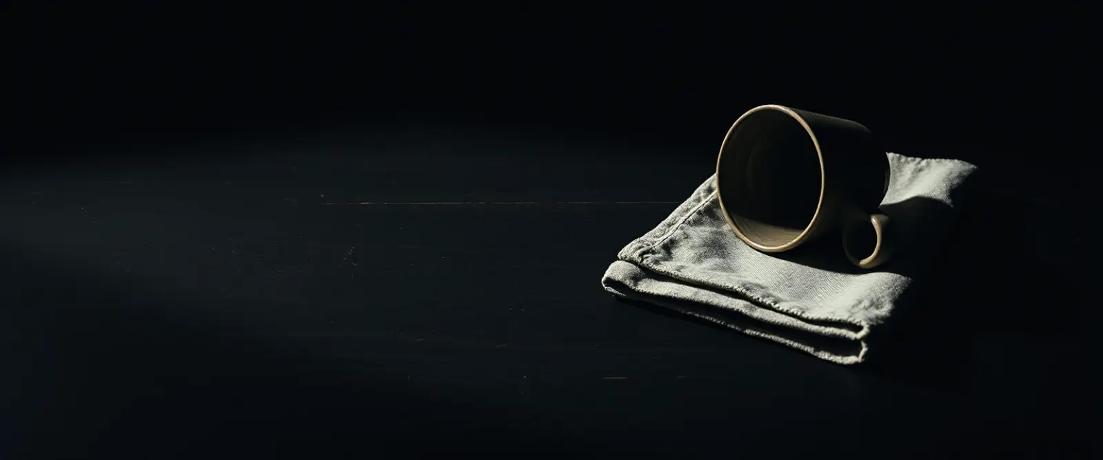 An empty dark cup turned upside down on a folded linen cloth, a dry ring on the wood beside it.