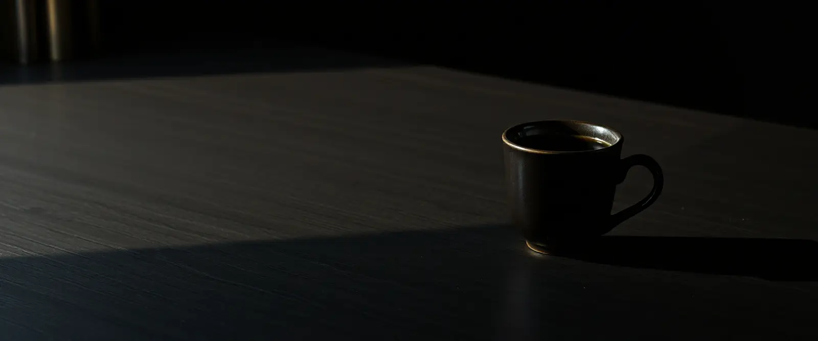 A single dark ceramic cup of black coffee on a wide table in a dim room.