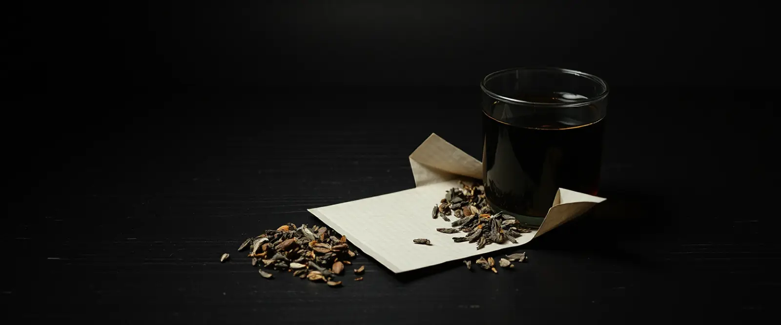 An open paper packet of loose-leaf tea spilling onto a dark table, a glass of brewed tea behind it.