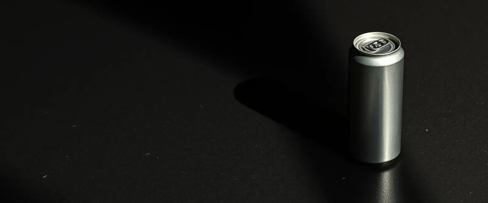 A plain unlabelled aluminium can on a dark concrete surface, its shadow long across the frame.