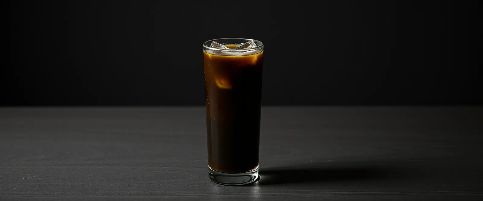 A tall glass of very dark iced coffee on a counter, beaded with condensation.