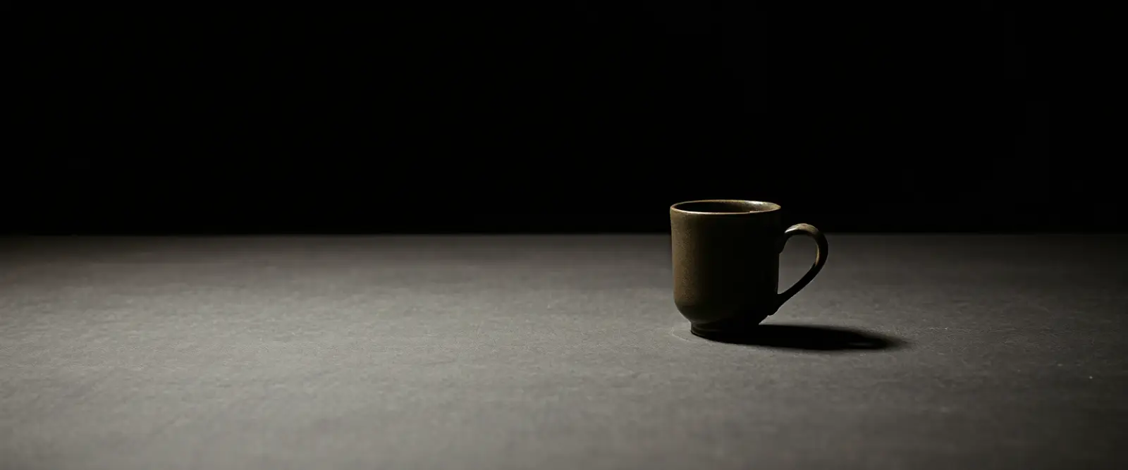A single dark ceramic cup of black coffee on a wide table in a dim room.