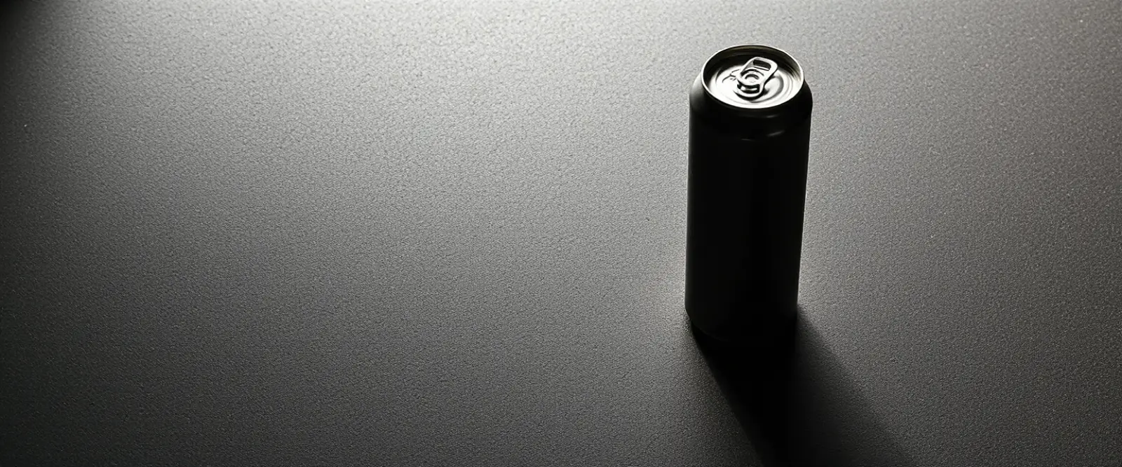 A plain unlabelled aluminium can on a dark concrete surface, its shadow long across the frame.