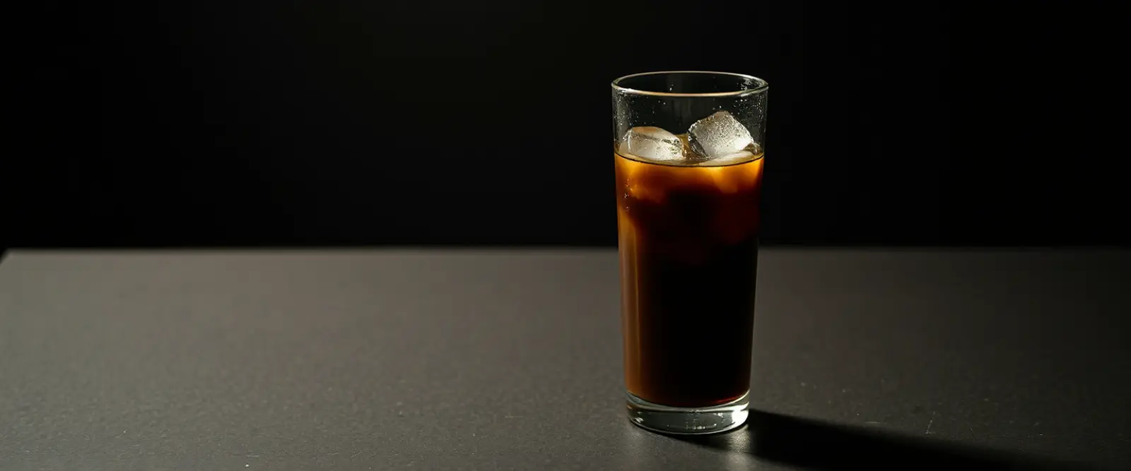 A tall glass of very dark iced coffee on a counter, beaded with condensation.
