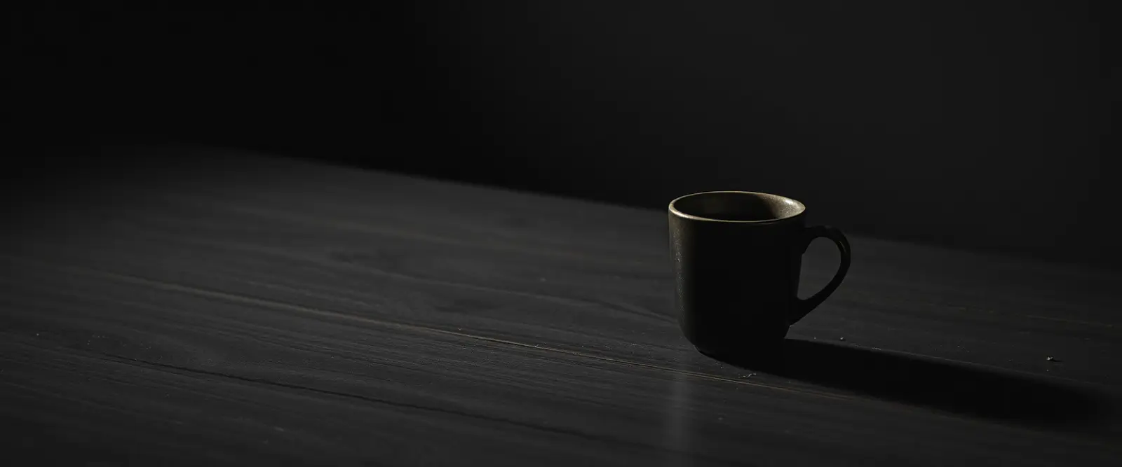 A single dark ceramic cup of black coffee on a wide table in a dim room.