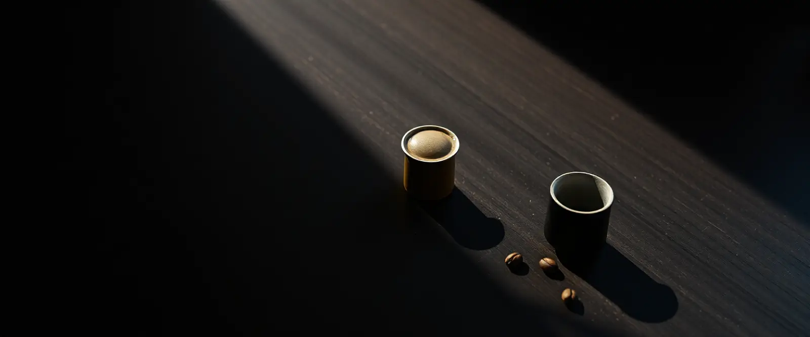 Three coffee capsules and a small dark cup on a table, lit from one side.