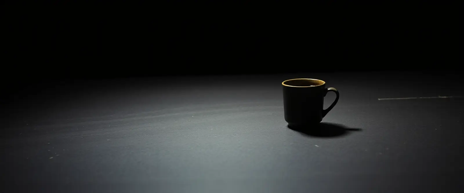 A single dark ceramic cup of black coffee on a wide table in a dim room.
