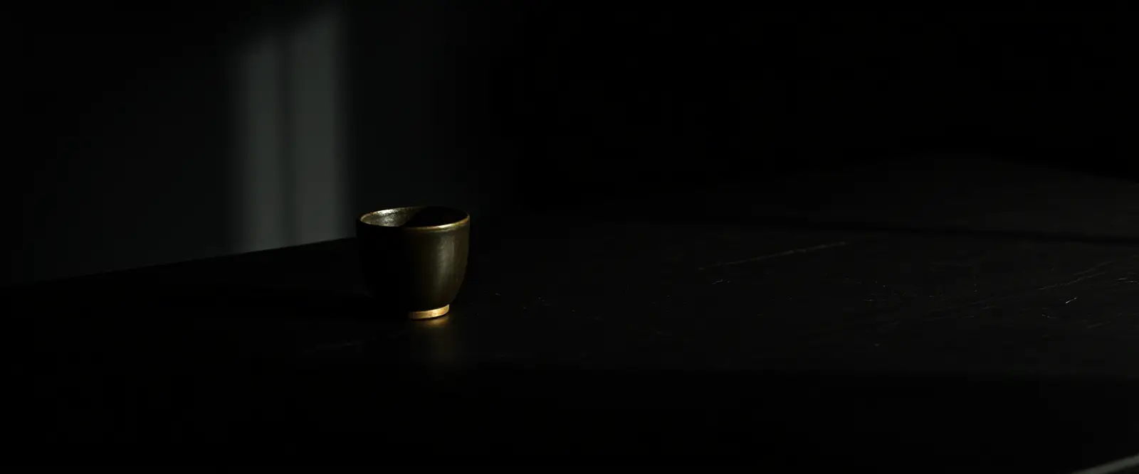 A single dark ceramic cup of black coffee on a wide table in a dim room.