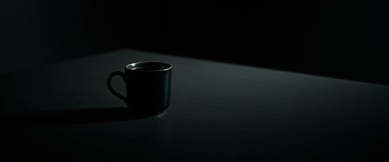 A single dark ceramic cup of black coffee on a wide table in a dim room.