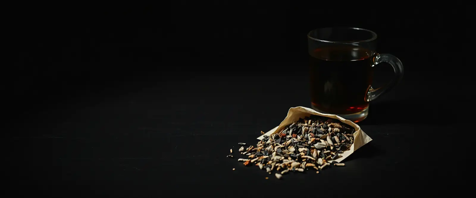 An open paper packet of loose-leaf tea spilling onto a dark table, a glass of brewed tea behind it.