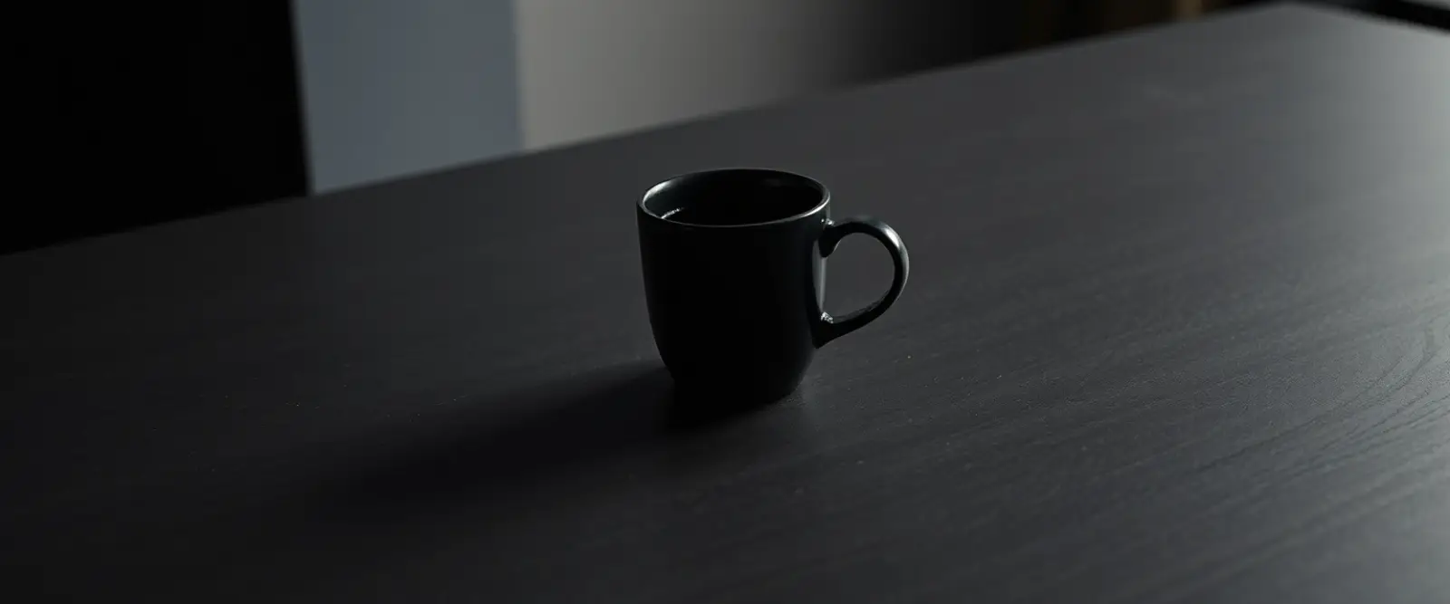 A single dark ceramic cup of black coffee on a wide table in a dim room.